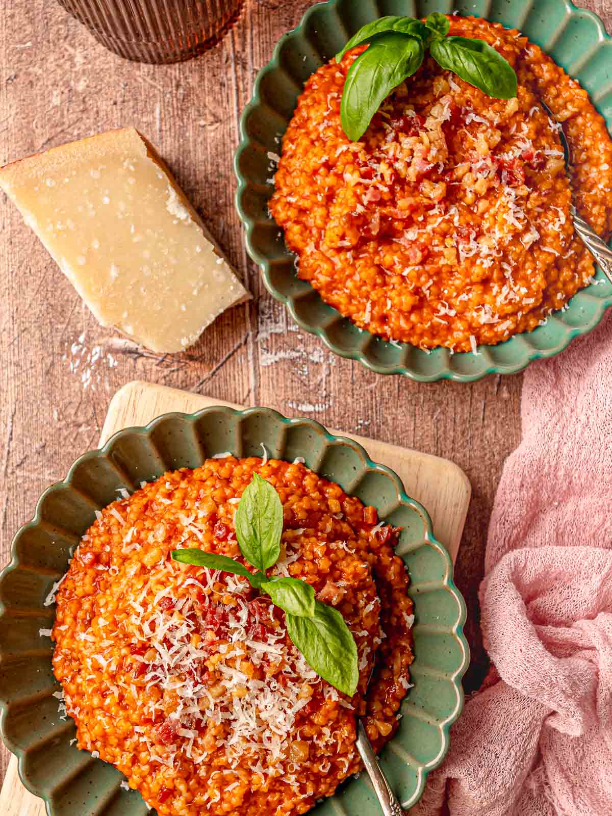 Two bowls of creamy tomato pastina with guanciale garnished with grated Pecorino Romano and fresh basil leaves in green scalloped-edge bowls, with a wedge of Pecorino cheese and a pink napkin alongside.