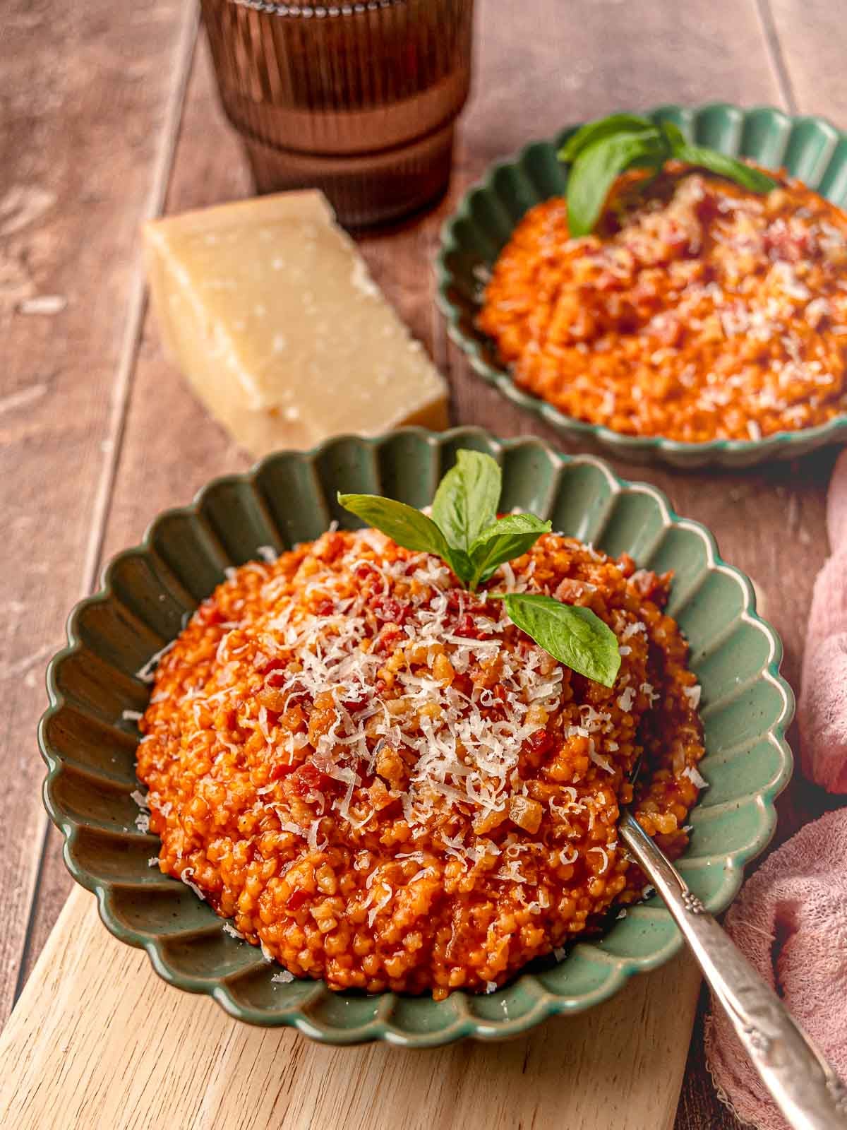 Two servings of tomato pastina with guanciale in green scalloped-edge bowls, topped with grated Pecorino and basil, with a wedge of cheese and pink napkin nearby.