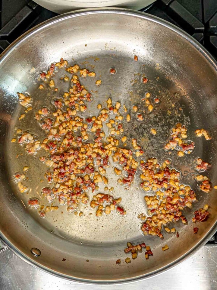 Diced guanciale rendering in a stainless steel skillet, releasing fat as it cooks.