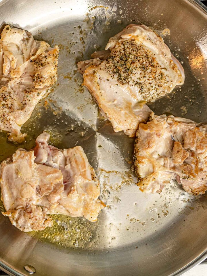 Seasoned chicken thighs searing in stainless steel skillet with golden-brown edges.