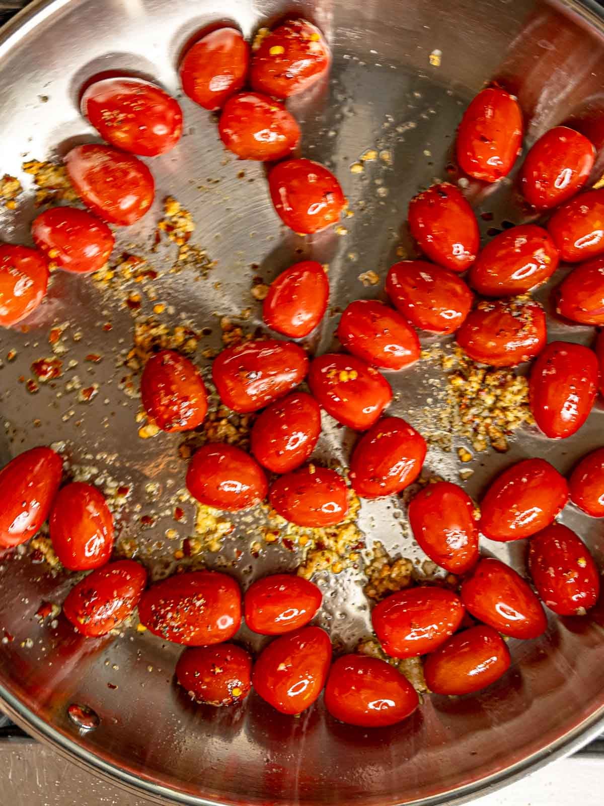 Cherry tomatoes cooking in olive oil with garlic and red pepper flakes in stainless steel skillet.