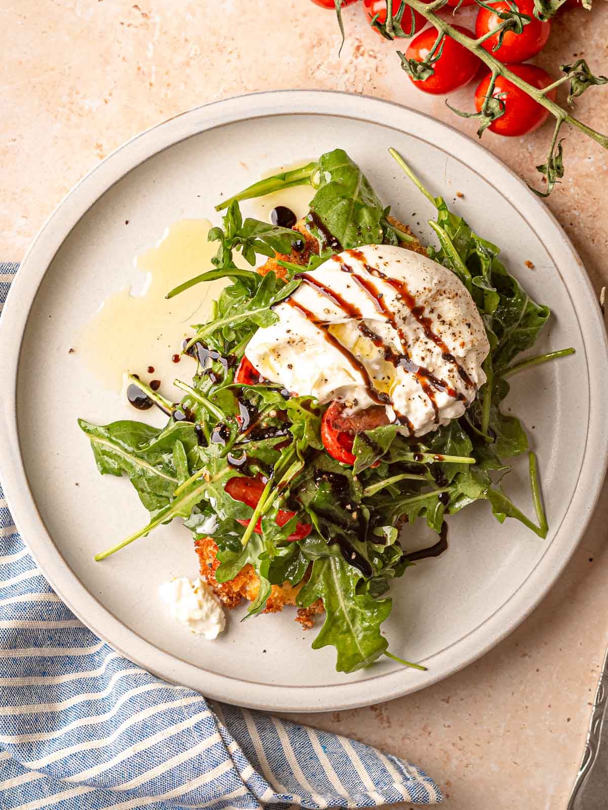 Plated chicken milanese with arugula, cherry tomatoes, burrata, and balsamic glaze with striped napkin.