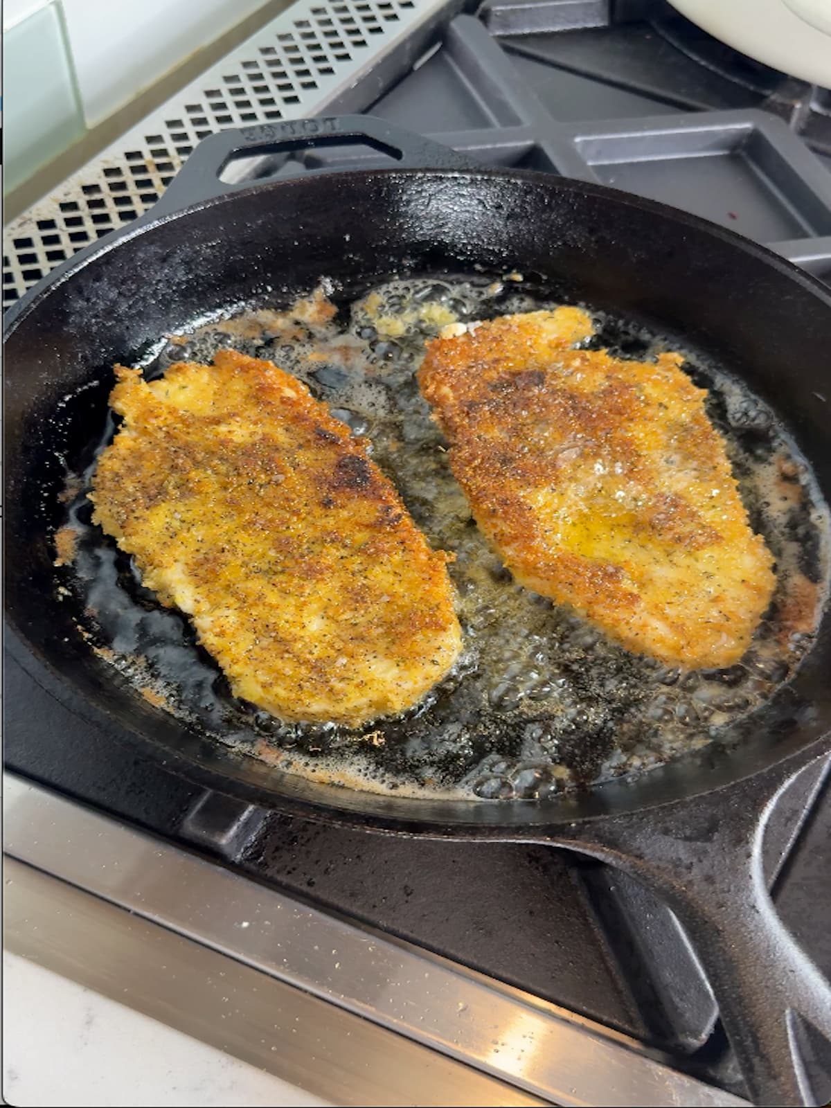 Two breaded chicken cutlets frying in bubbling oil in black cast iron skillet.