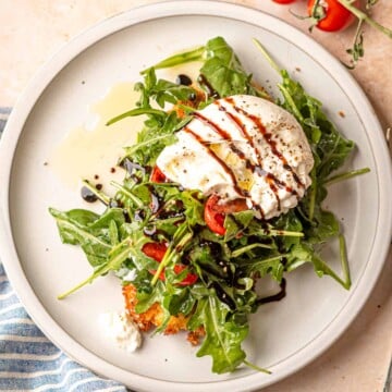 Chicken milanese topped with dressed arugula, cherry tomatoes, burrata, and balsamic glaze on white plate.
