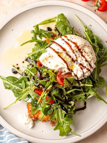 Chicken milanese topped with dressed arugula, cherry tomatoes, burrata, and balsamic glaze on white plate.