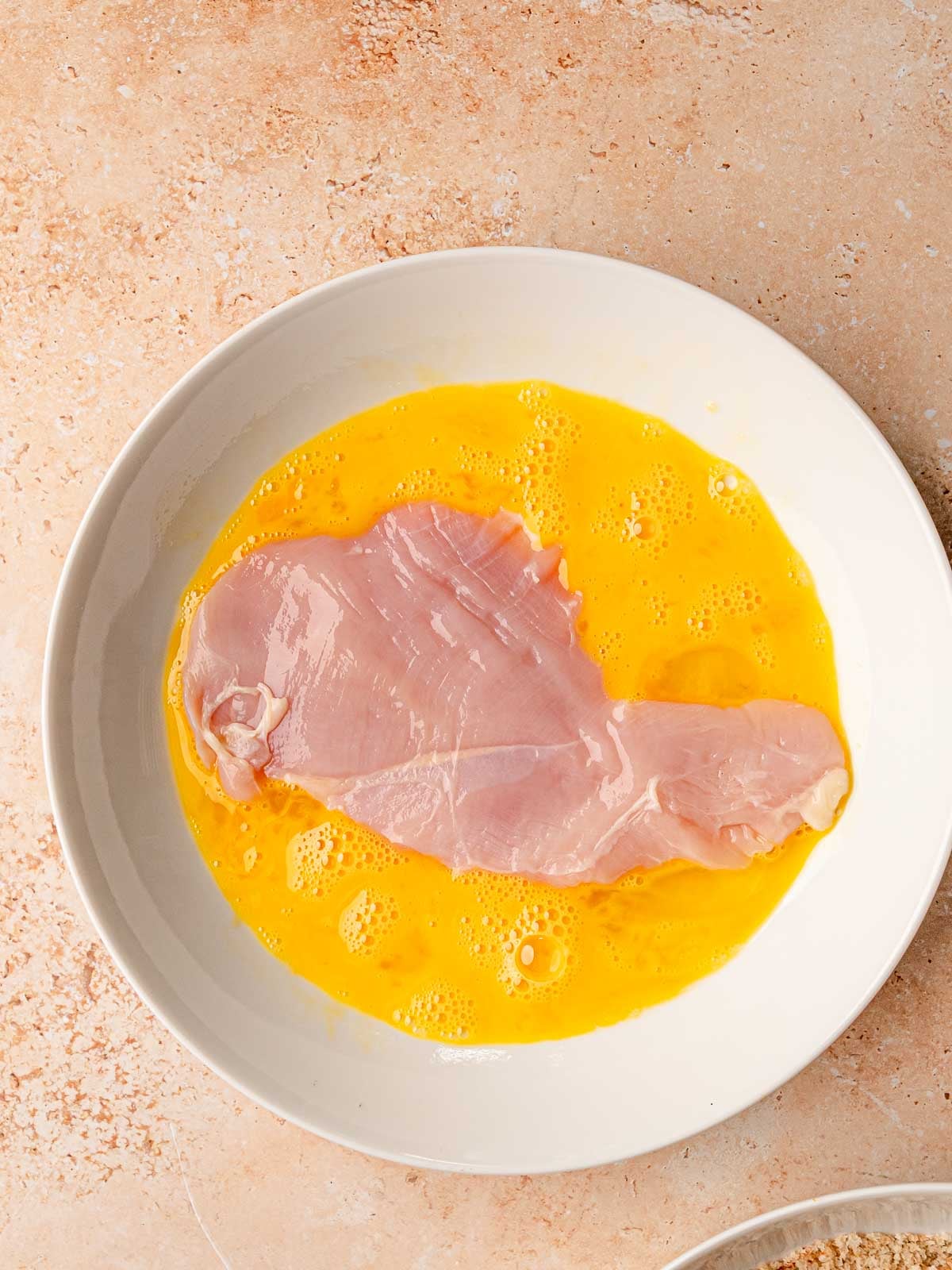 Pounded chicken cutlet soaking in beaten eggs in white bowl.