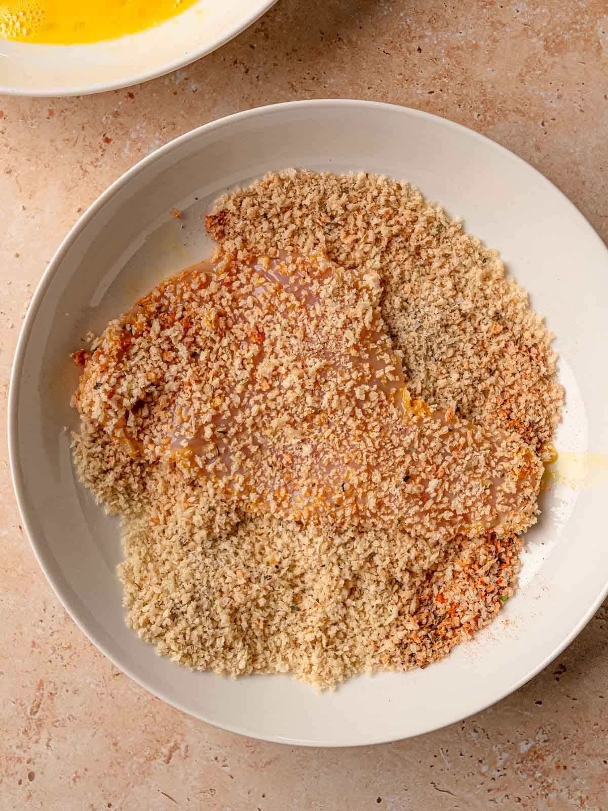 Egg-coated chicken cutlet pressed into panko breadcrumb mixture in white bowl.
