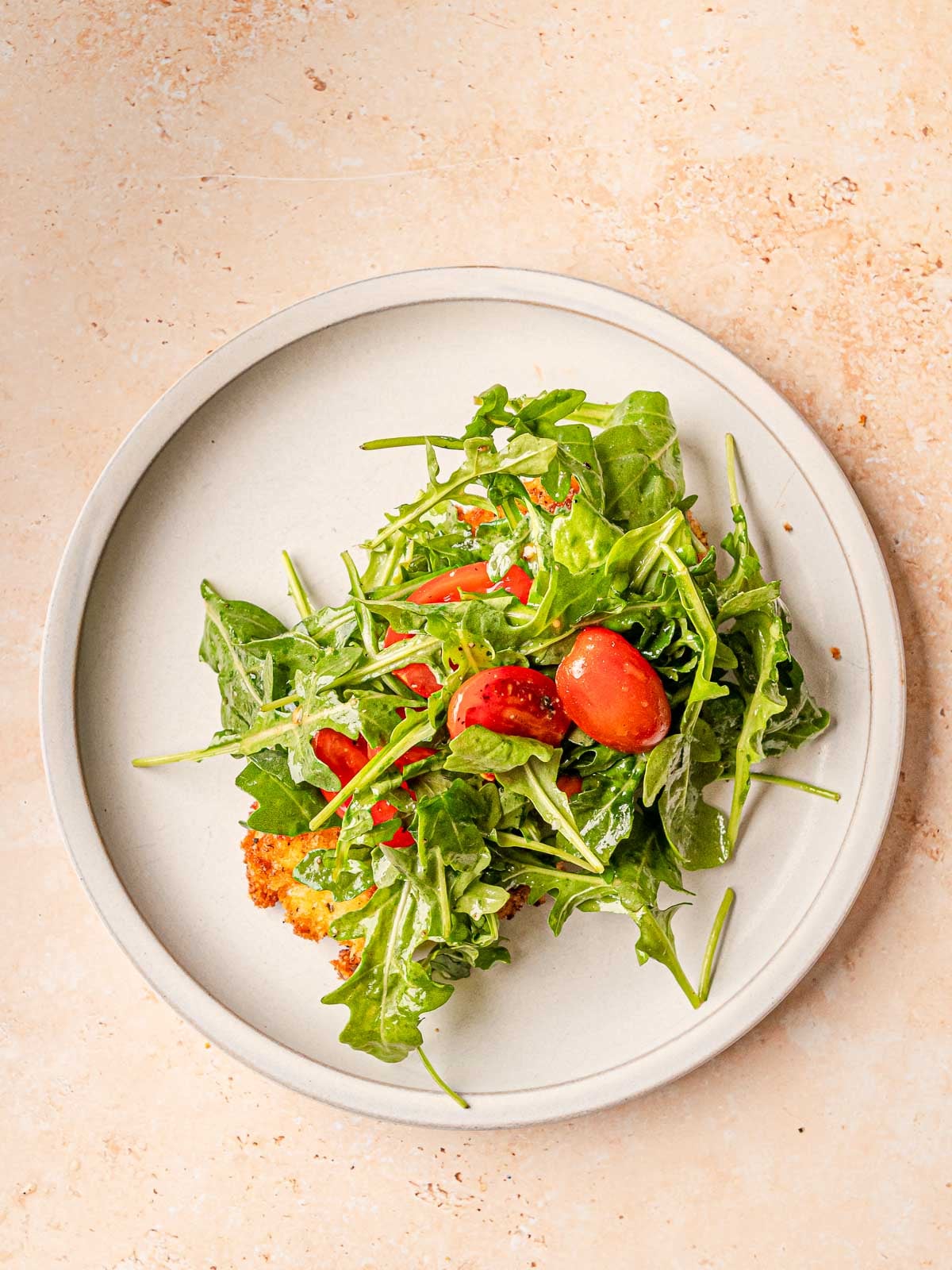 Crispy chicken milanese topped with arugula salad and cherry tomatoes on white plate.