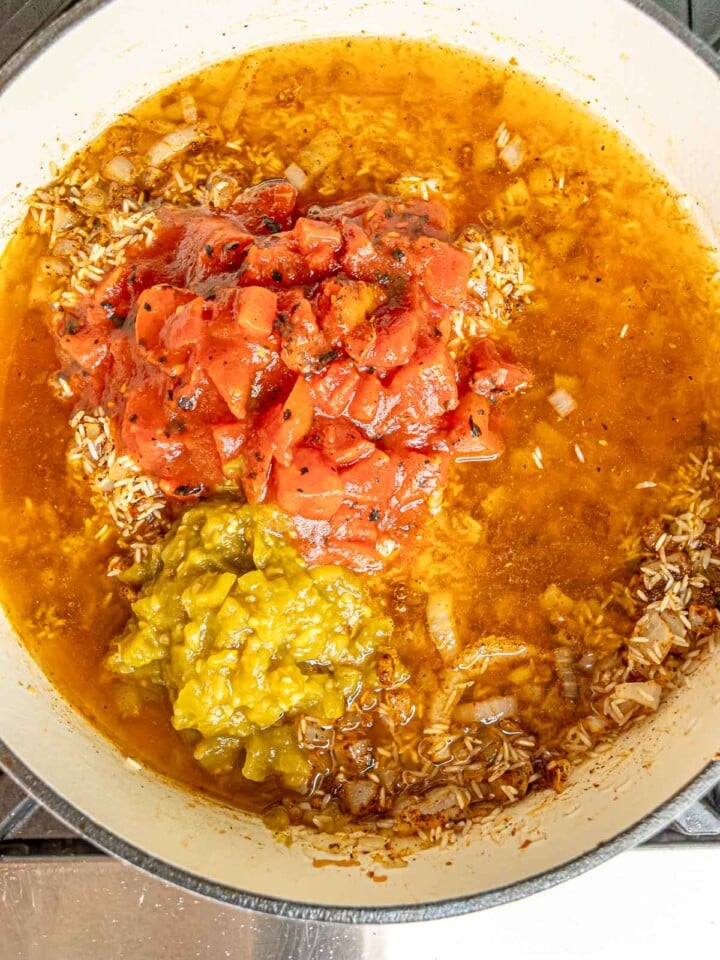 White skillet filled with broth, onions, rice, diced tomatoes, green chiles, and corn before stirring.