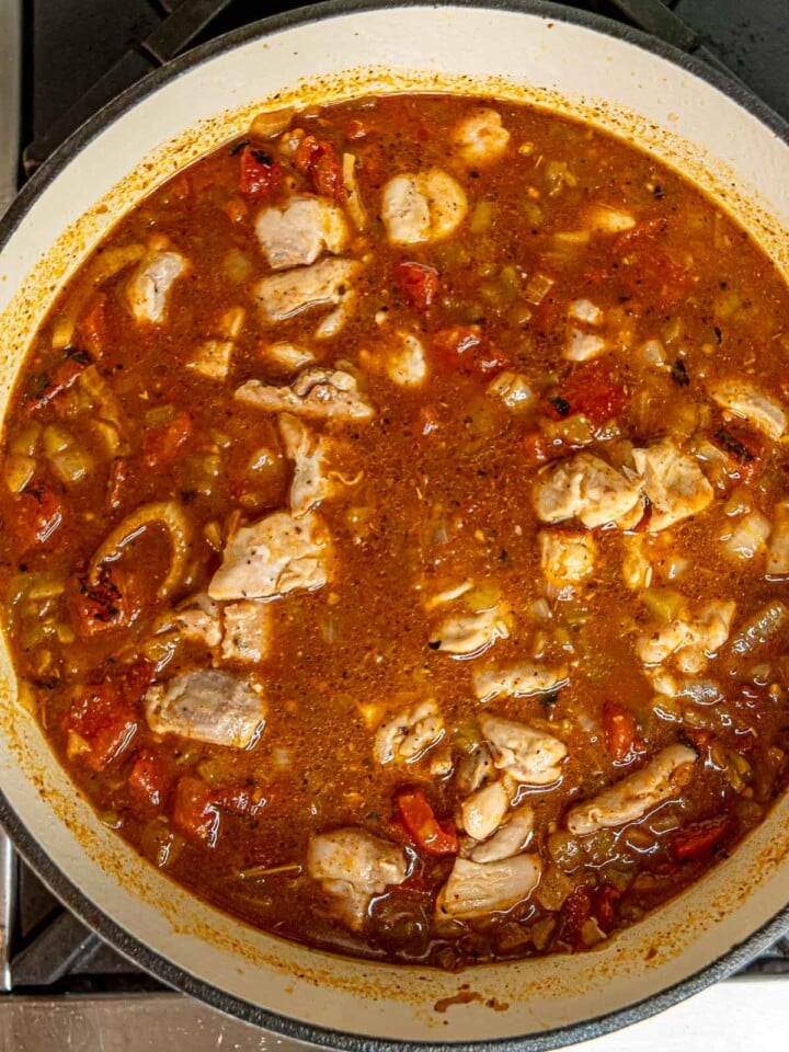 Bite-sized chicken pieces cooking in spiced broth with diced tomatoes and green chiles in white skillet.