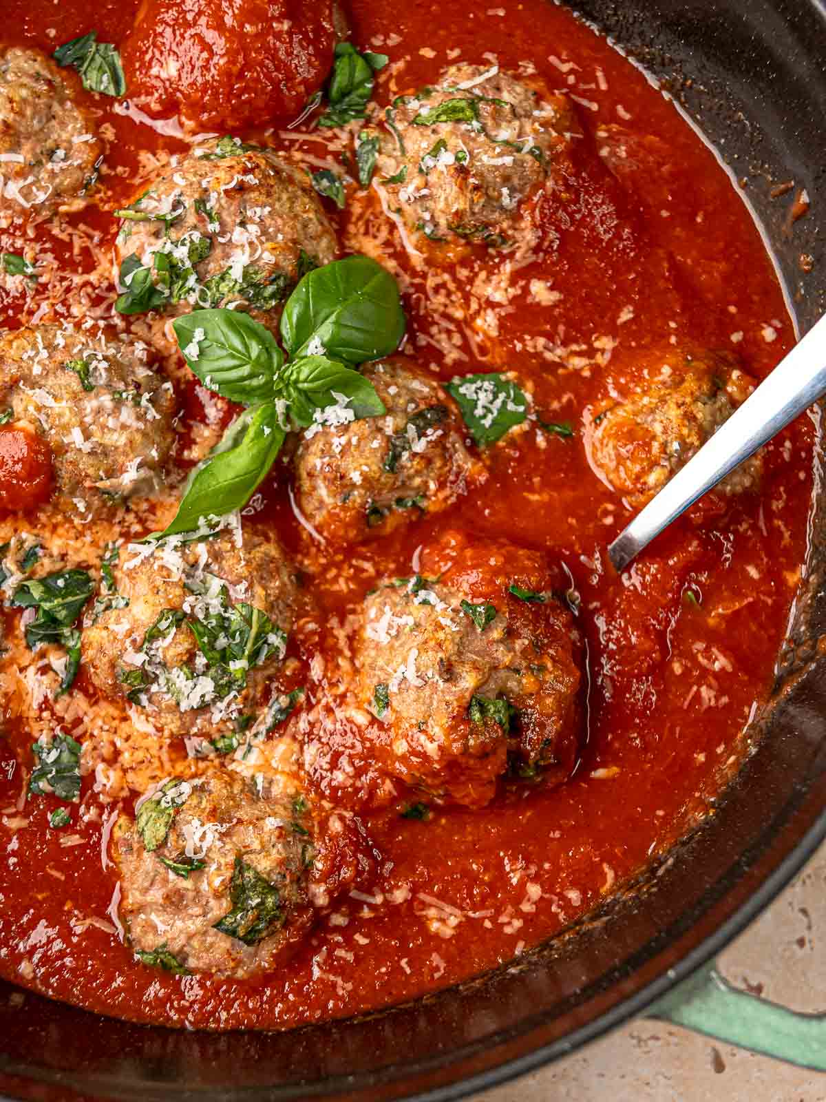 Serving spoon lifting turkey spinach meatball from cast iron skillet with tomato sauce.