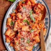 Rigatoni with pancetta arrabbiata sauce is served on a blue and white platter with grated Parmesan and fresh basil.