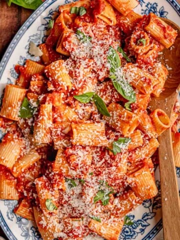 Rigatoni with pancetta arrabbiata sauce is served on a blue and white platter with grated Parmesan and fresh basil.