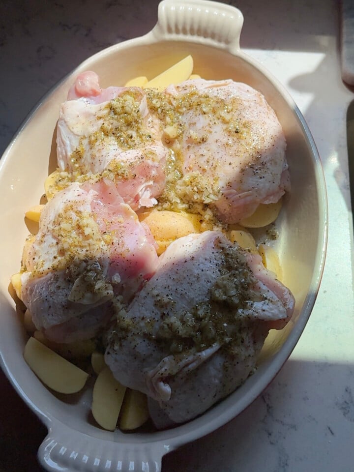 Raw chicken thighs and potatoes are coated with a garlic herb marinade in a baking dish before cooking.