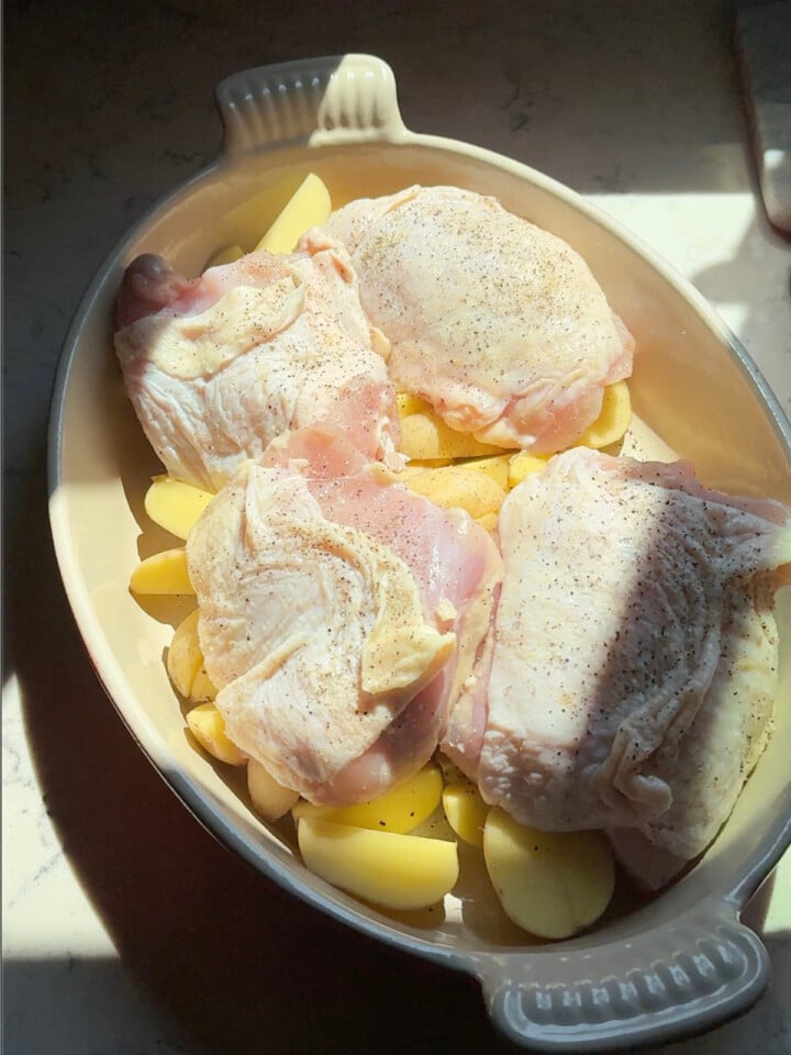 Raw seasoned chicken thighs are arranged over sliced baby potatoes in a baking dish.