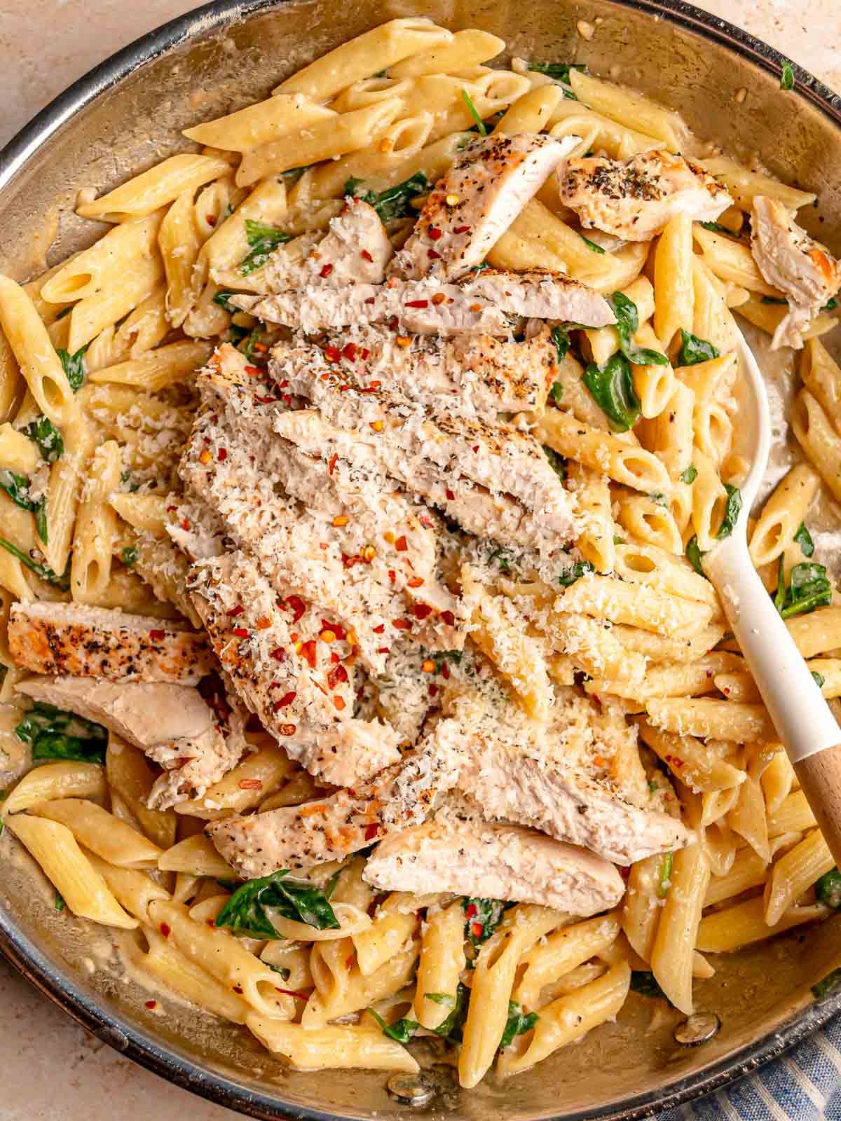 Creamy garlic Parmesan chicken pasta with penne, spinach and sliced seasoned chicken topped with red pepper flakes and grated cheese.