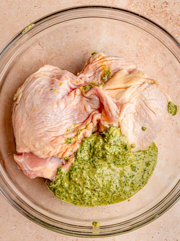 Bone-in chicken thighs with green herb and yogurt marinade in a glass bowl.