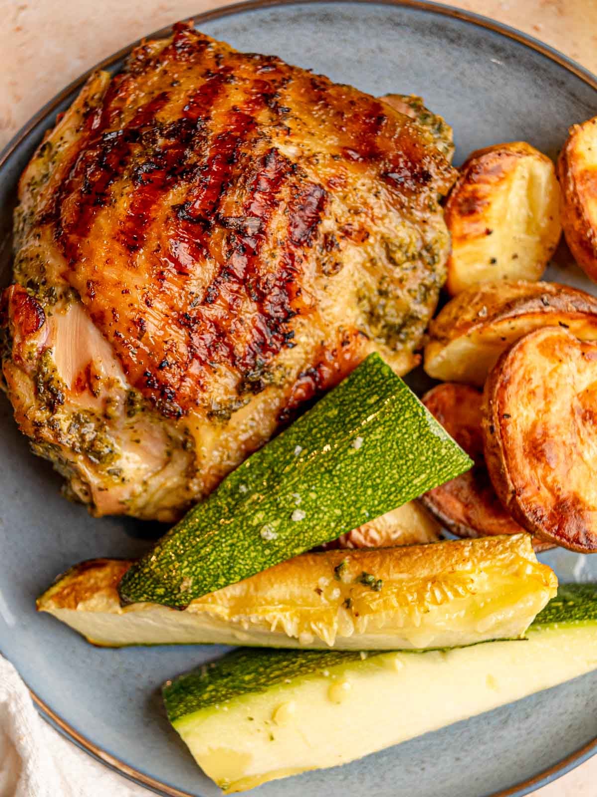 Grilled chicken thigh with golden, charred skin served alongside roasted potatoes and zucchini.