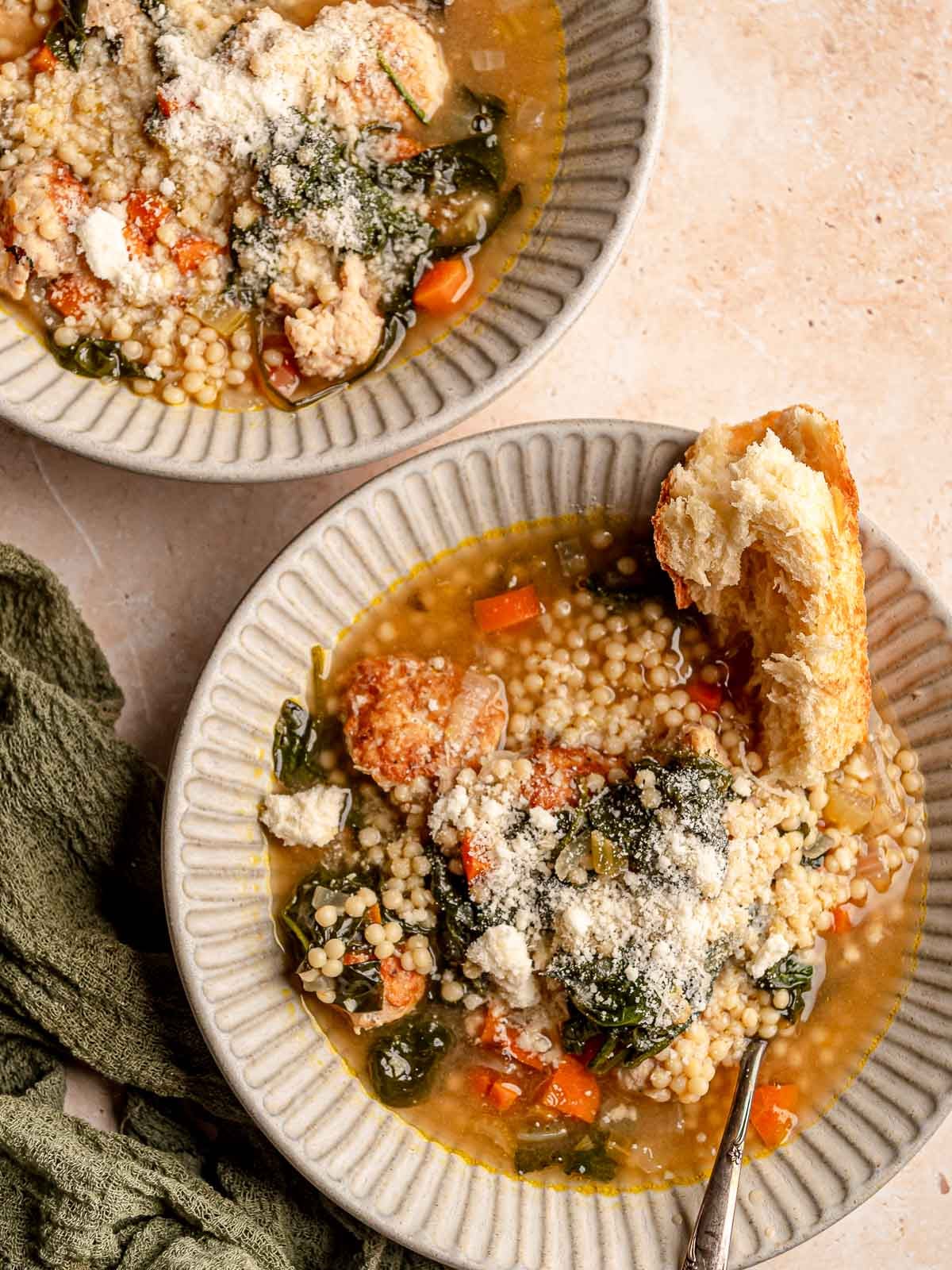 Two bowls of Italian wedding soup with chicken meatballs, acini di pepe, spinach, vegetables, and Parmesan are served with crusty bread.