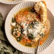 A bowl of Italian wedding soup with chicken meatballs, pasta, spinach, and vegetables is topped with grated Parmesan and served with bread.
