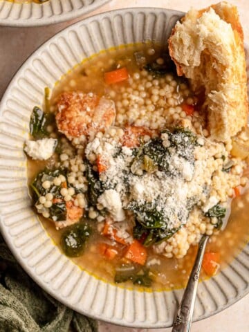 A bowl of Italian wedding soup with chicken meatballs, pasta, spinach, and vegetables is topped with grated Parmesan and served with bread.