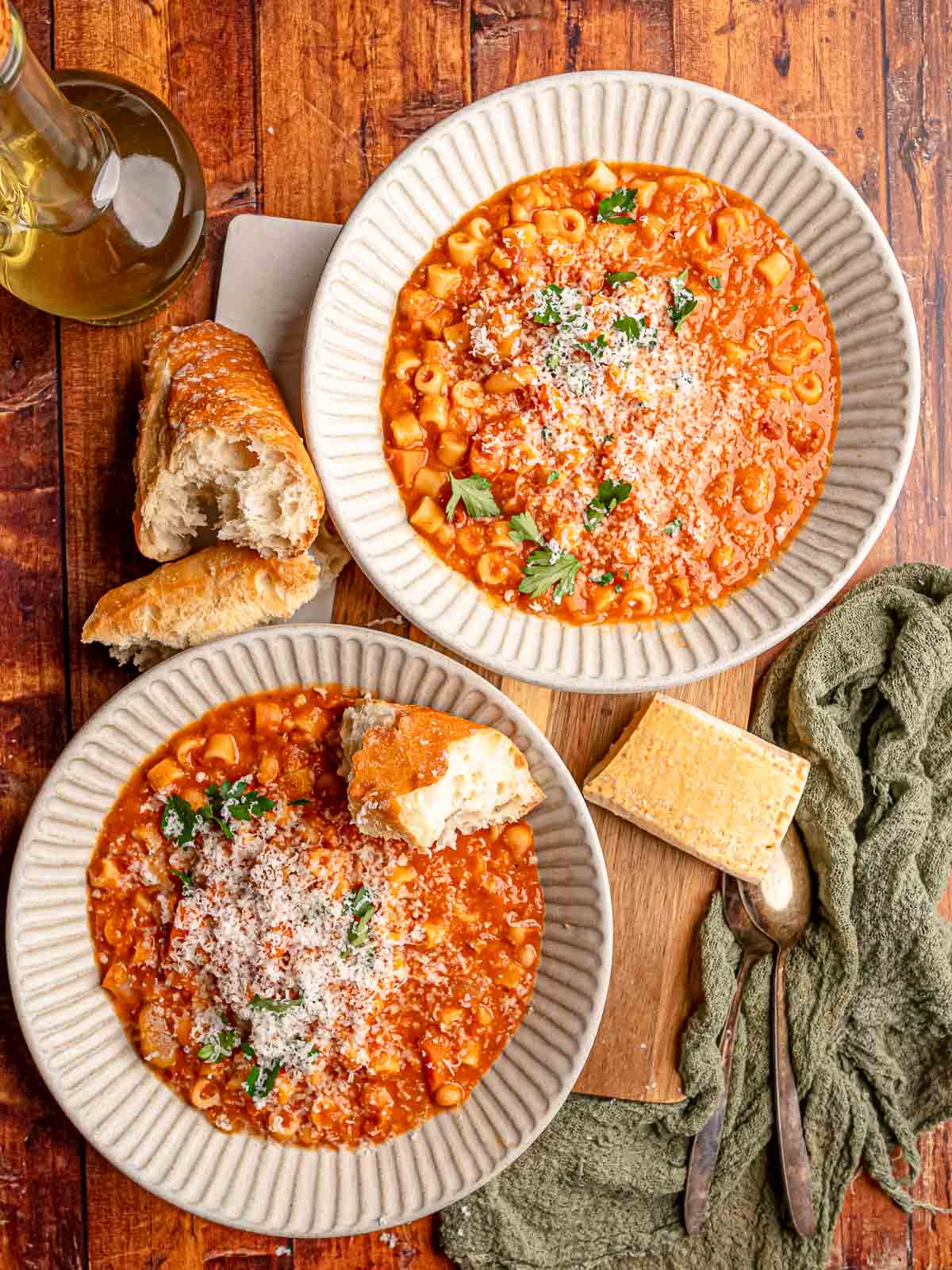 Two bowls of pasta e fagioli with beans, ditalini, Parmesan, and parsley are served with crusty bread on a wooden table.
