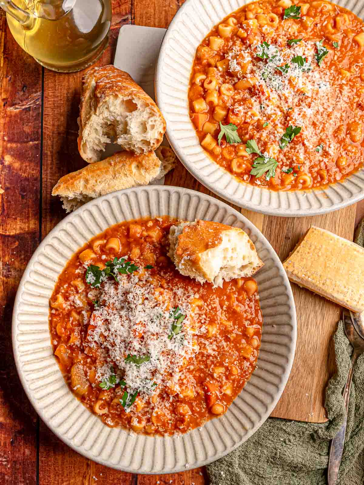 Two bowls of pasta e fagioli with ditalini and cannellini beans are topped with Parmesan and parsley and served with crusty bread.