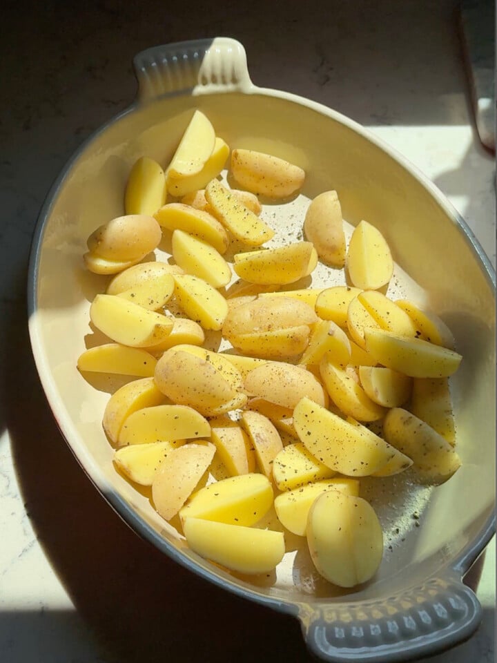 Quartered baby potatoes are seasoned with salt and pepper in a baking dish before the chicken is added.