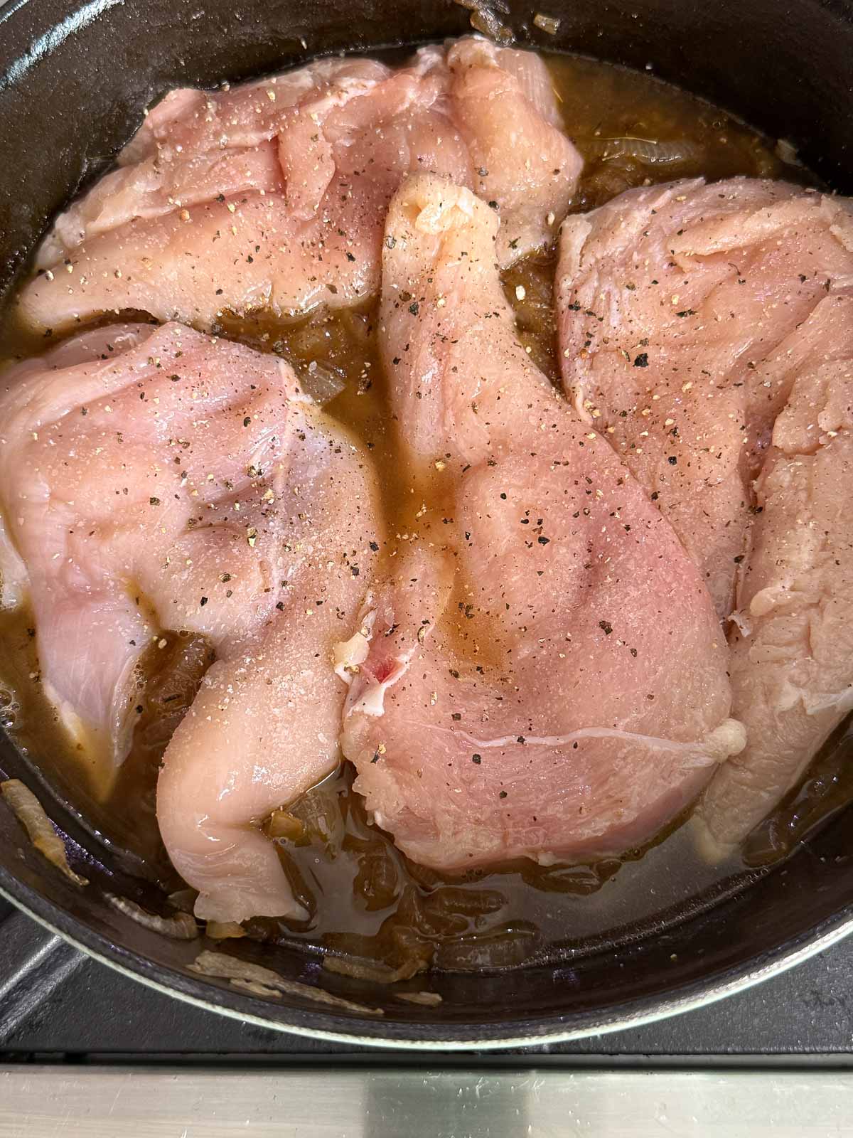 Seasoned raw chicken breasts nestled into caramelized onions and broth in a skillet.