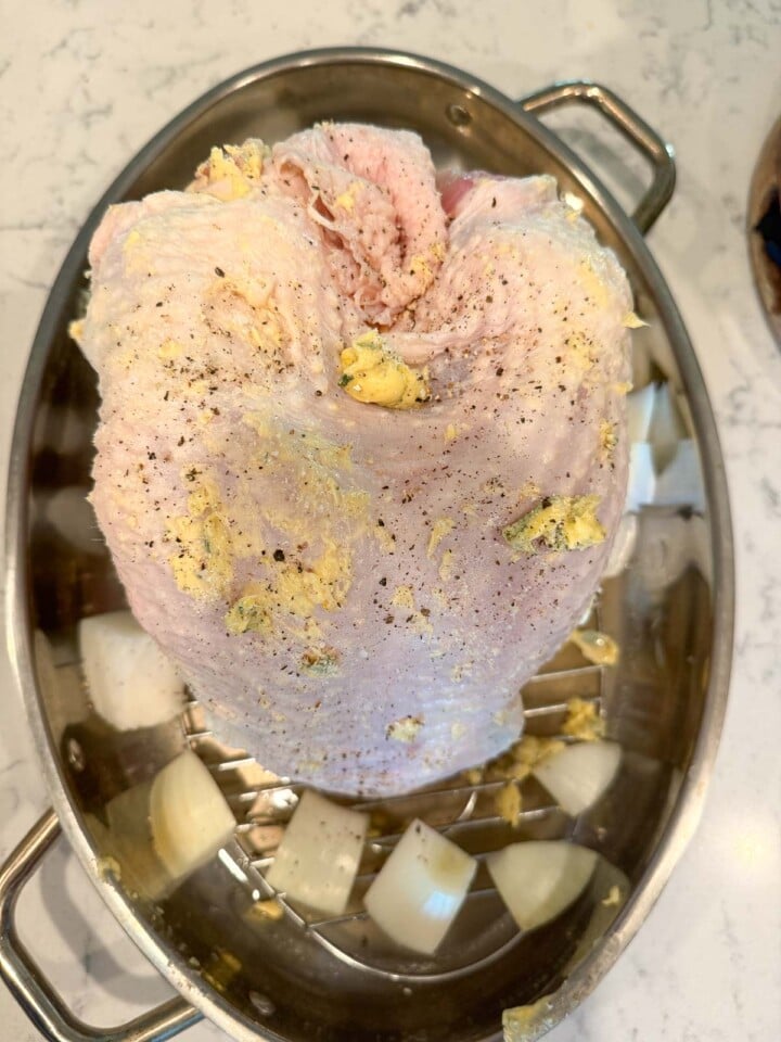 Raw turkey breast coated with herb butter on a roasting rack over chopped onions.